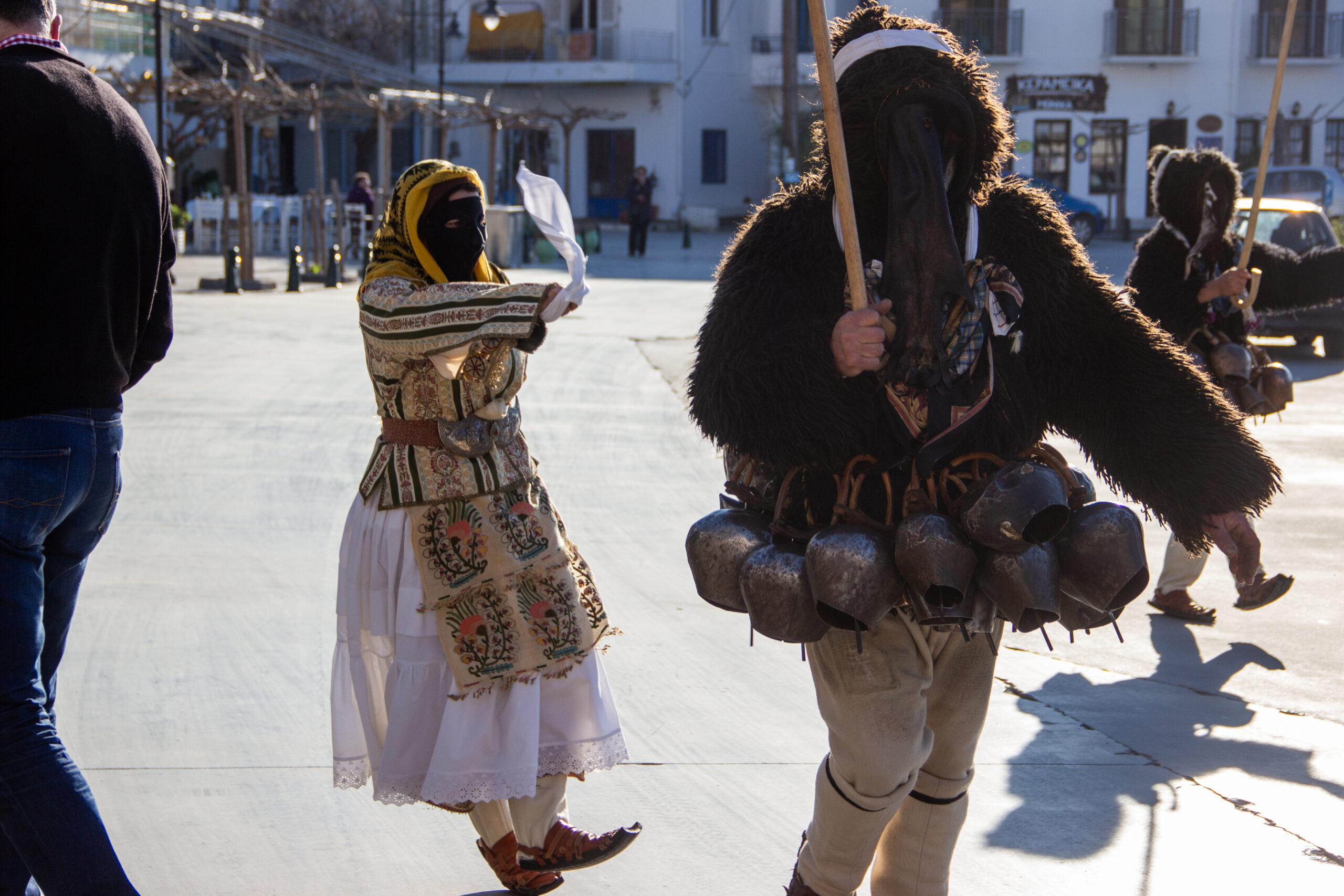 A Skyros masked goatman wearing goatbells and a masked woman in yellow and white dress waving a white cloth perform in a square.