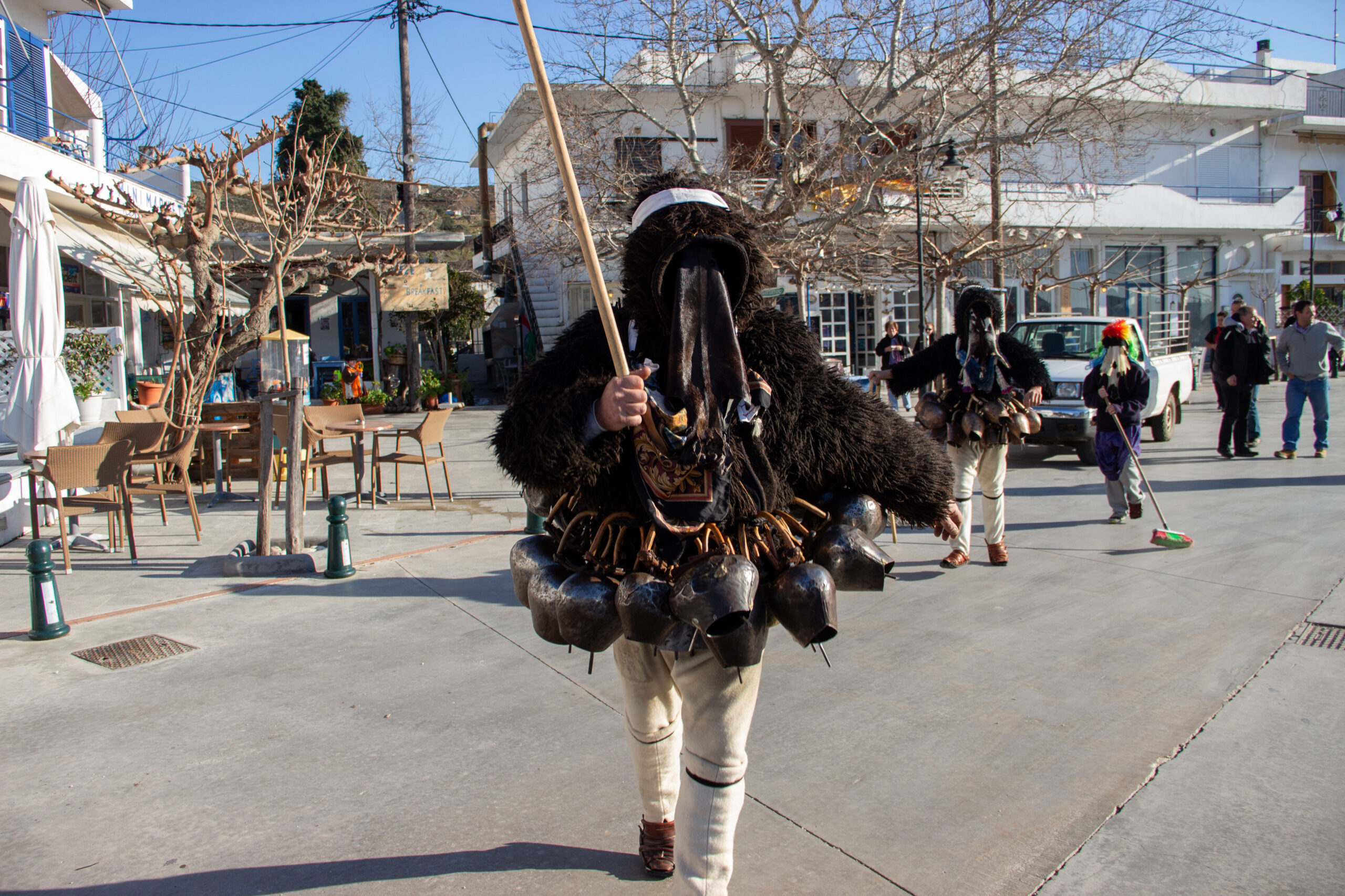 Close-up of a Skyros Carnival Geros, wearing a black furry jacket with a large belt of bells, a goatskin face mask and carrying a wooden stick.