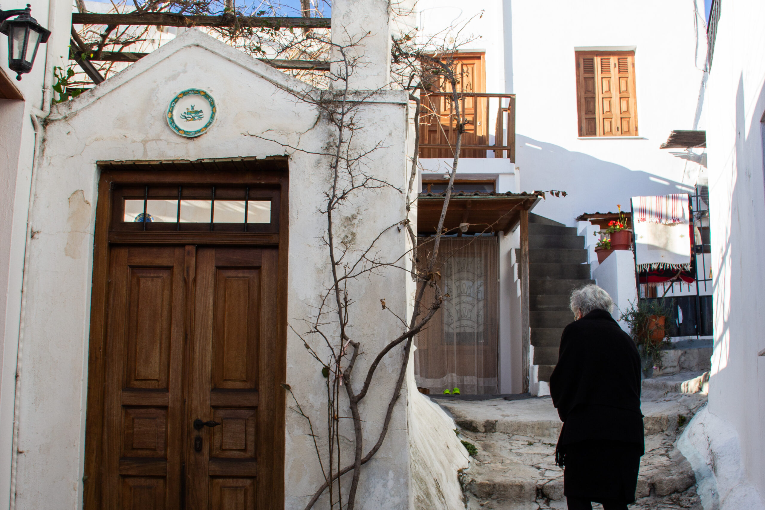 A woman in a black coat walks through a tiny street in Skyros Chora, past white houses with dark wooden doors.