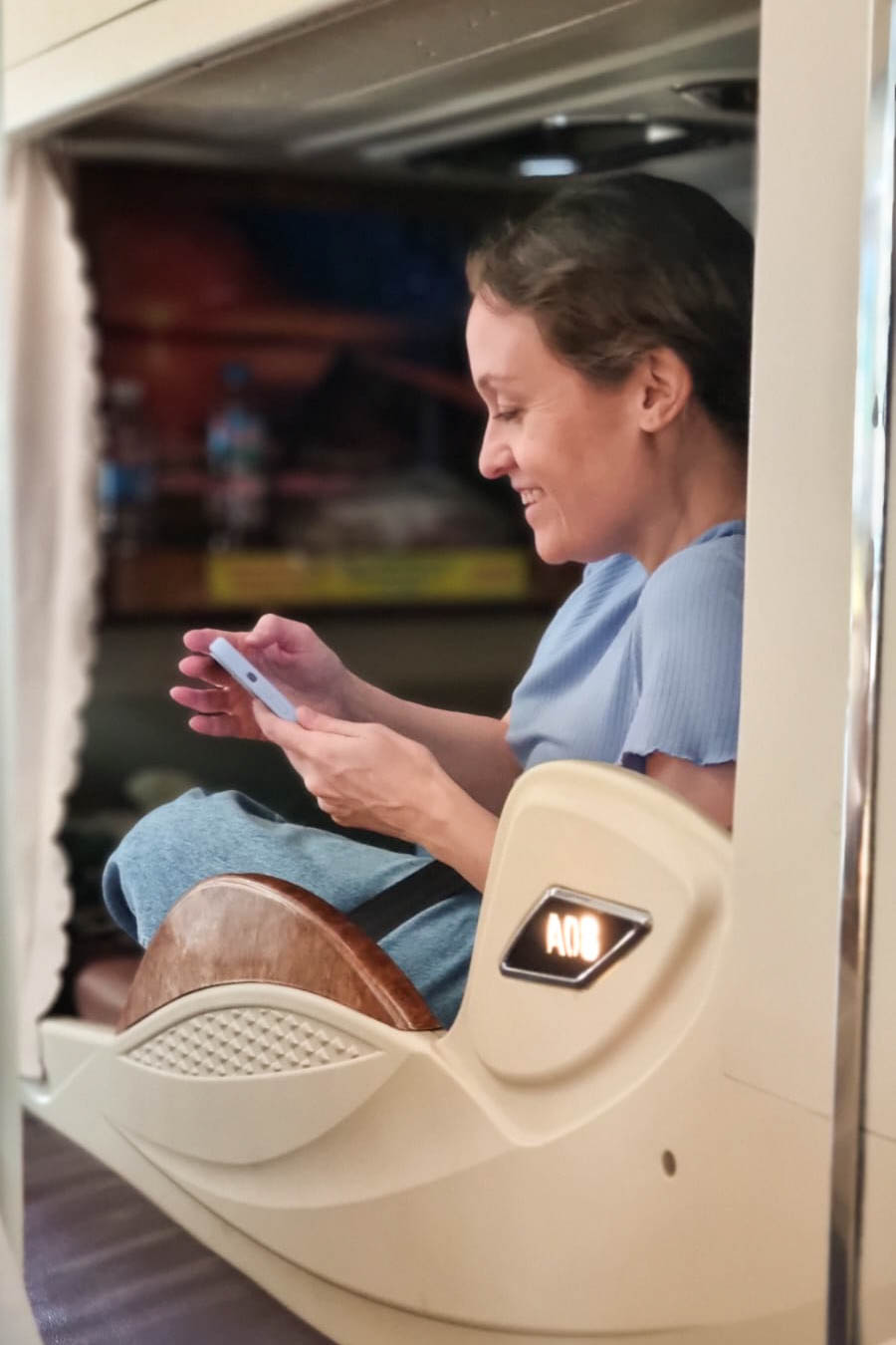 Smiling woman using a smartphone while sitting in a modern sleeper bus cabin with privacy curtains.