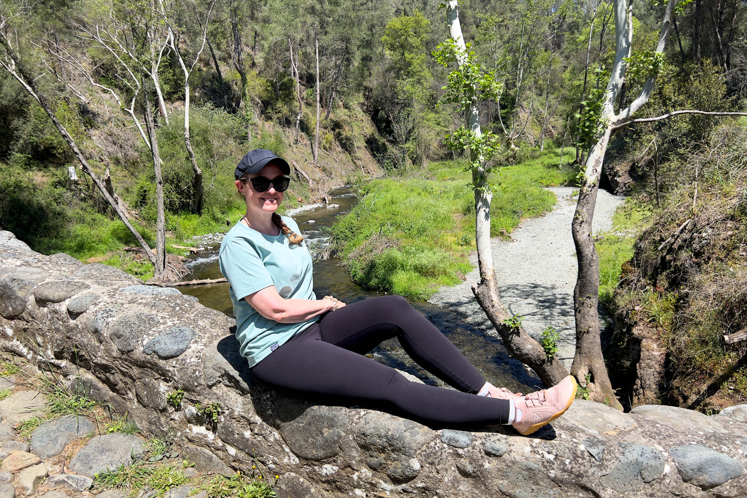 A woman in sunglasses and black cap, hiking clothing and pink trail shoes sitting on a stone wall overlooking a flowing creek in a green, wooded canyon.