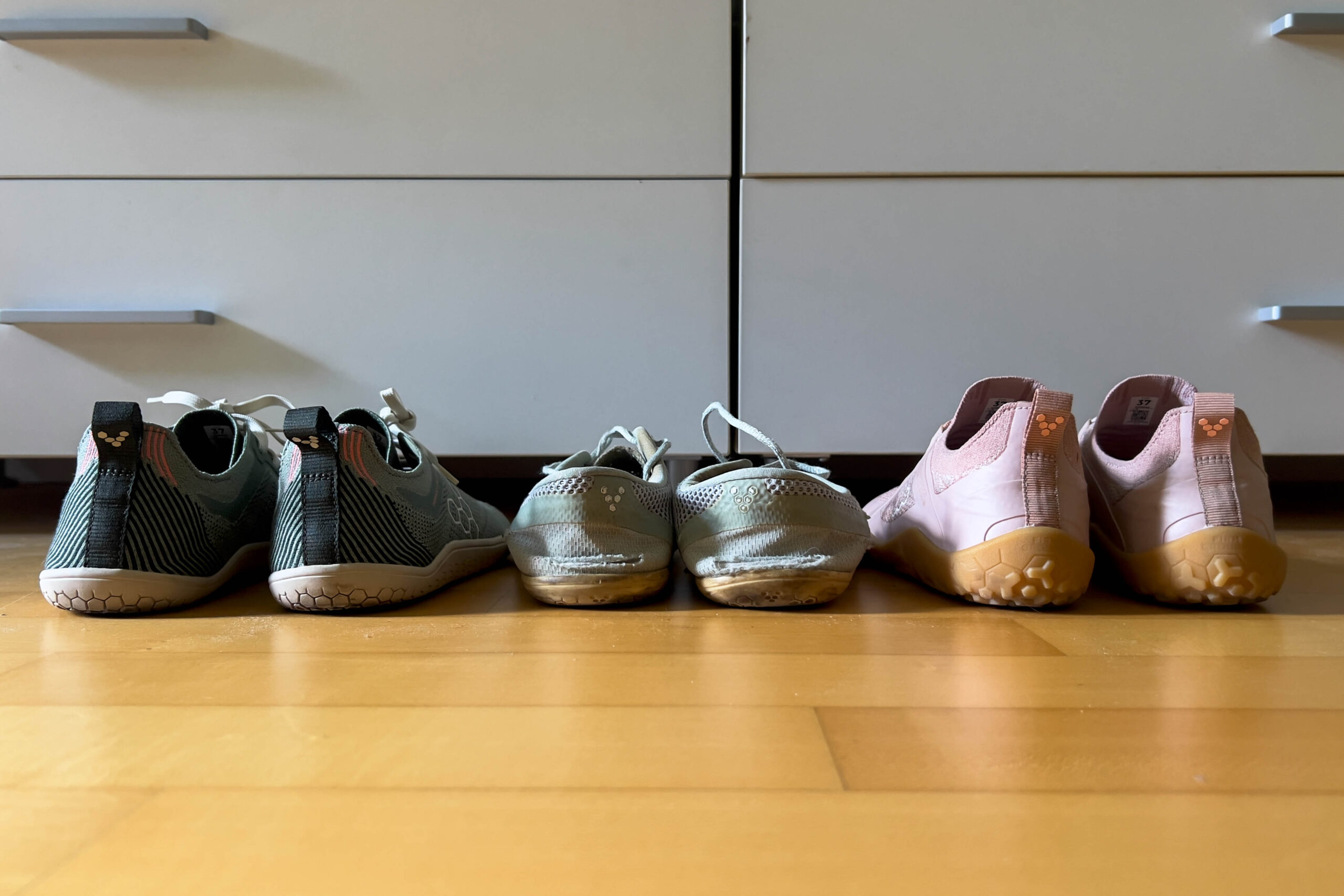 Back-view of three pairs of Vivobarefoot minimalist shoes lined up against a white dresser on a light wood floor.